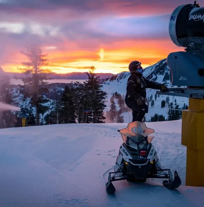 Person standing next to a snowmaking machine and a snowmobile on a snowy mountain at dawn