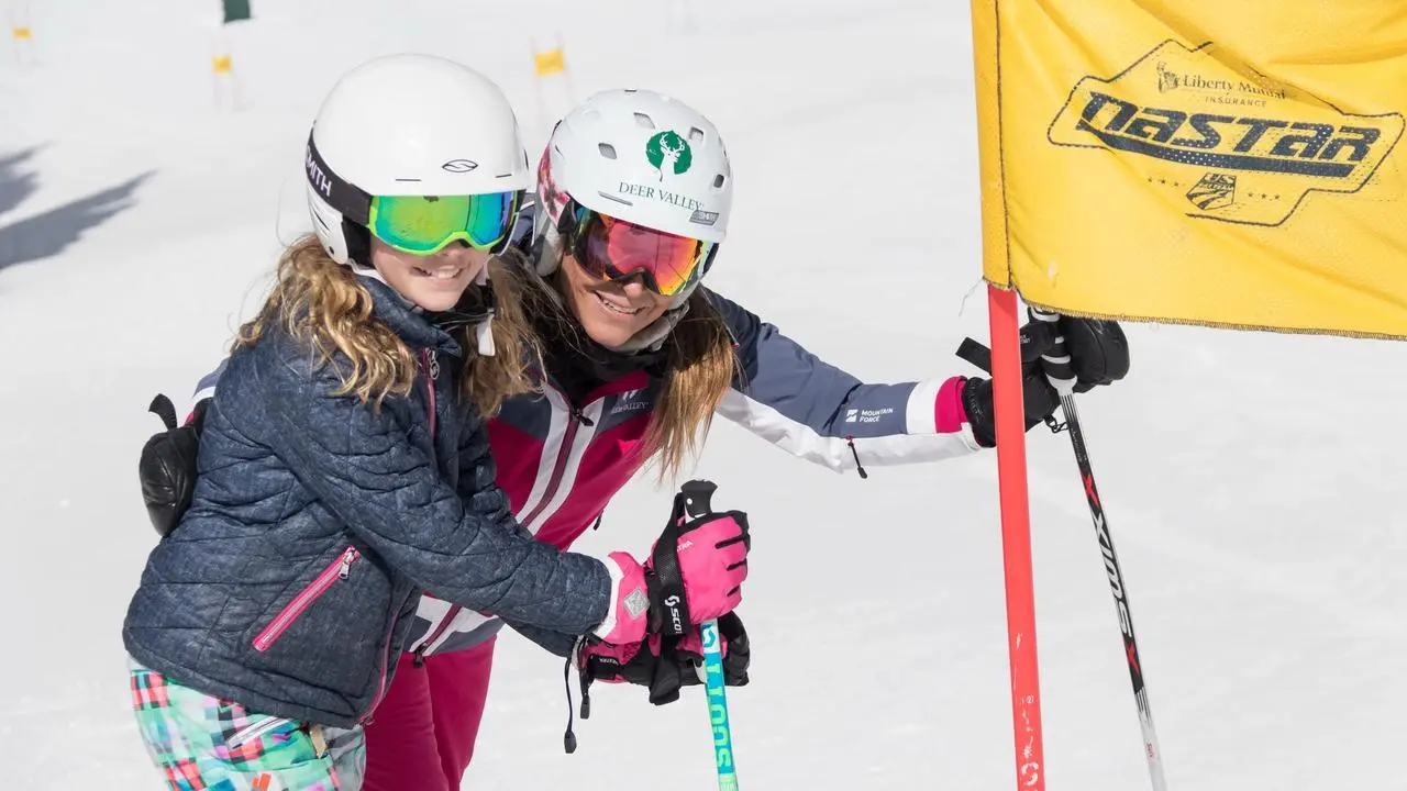 An adult and kid in ski gear standing next to a Nastar ski gate smiling at the camera