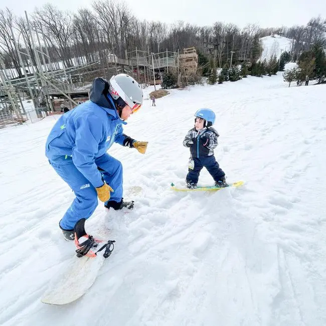 Adult dressed in winter gear riding a snowboard instructing a young child also dressed in winter gear strapped into a snowboard
