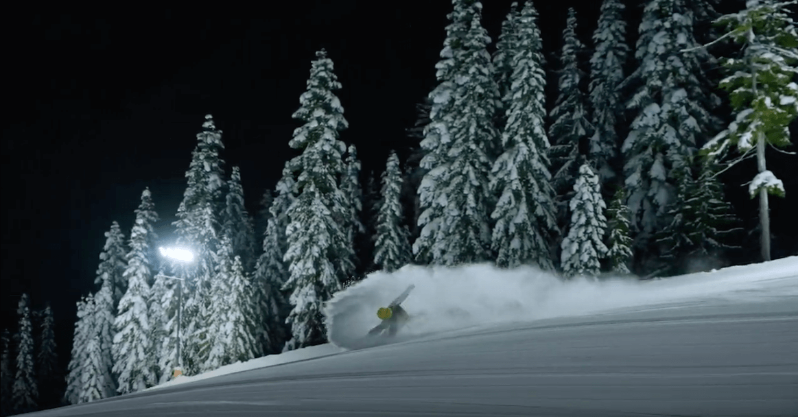 Person snowboarding down a snowy trail at night with lights shining on the run
